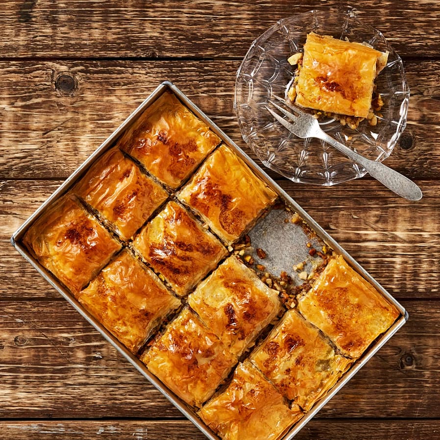 En plåt med skuren baklava med gyllene yta, bredvid en tallrik med en bit baklava och en gaffel.