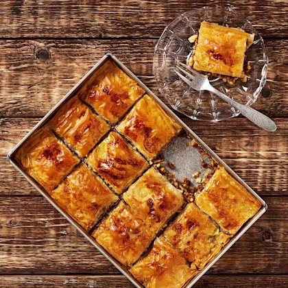 En plåt med skuren baklava med gyllene yta, bredvid en tallrik med en bit baklava och en gaffel.