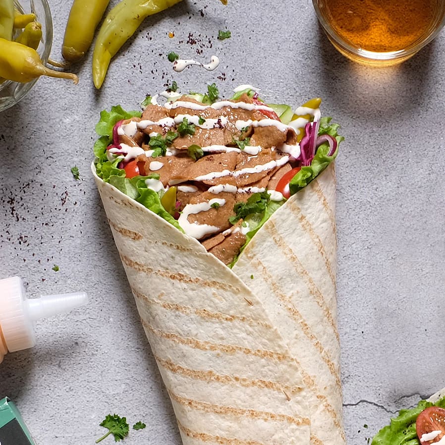 En fylld tortilla med kött, grönsaker och sås, omgiven av chilifrukter och en ölglas, på ett grått underlag.