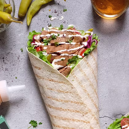 En fylld tortilla med kött, grönsaker och sås, omgiven av chilifrukter och en ölglas, på ett grått underlag.