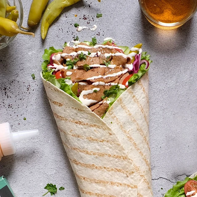 En fylld tortilla med kött, grönsaker och sås, omgiven av chilifrukter och en ölglas, på ett grått underlag.