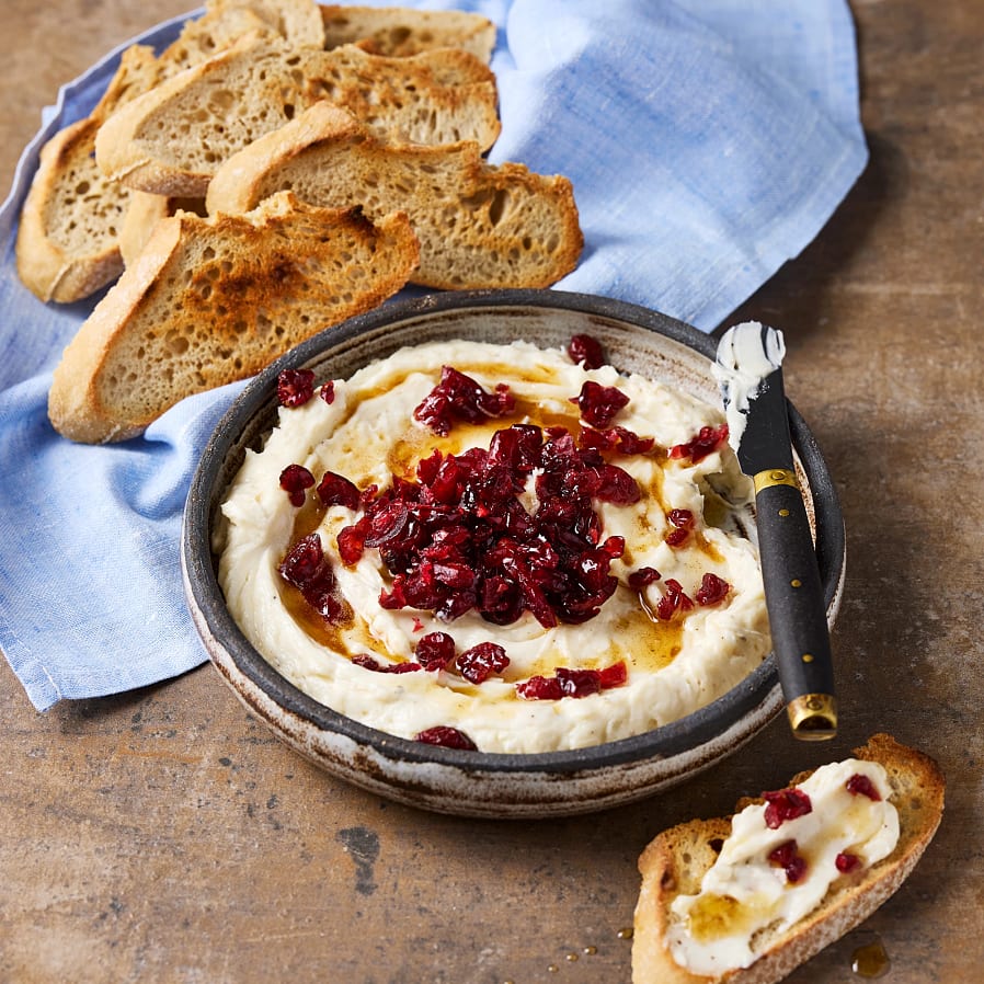 Getoströra med torkade tranbär och honung i en skål, serverad med rostade brödskivor på en duk.