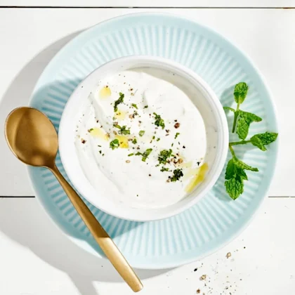 Myntasås i en vit skål som står på en turkos tallrik. Såsen är dekorerad med mynta, olja och svartpeppar. Till höger om skålen ligger en kvist mynta och till vänster om skålen ligger en sked.