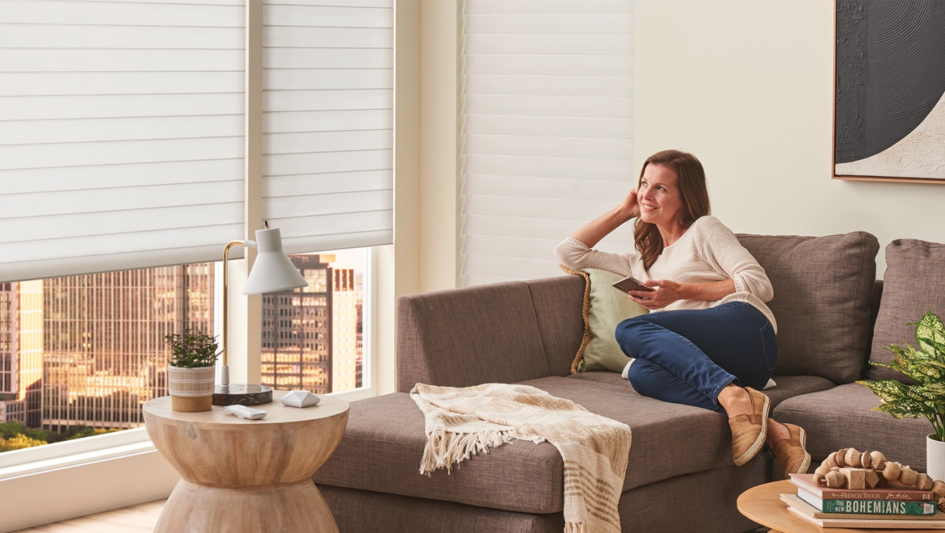 Woman sitting on her sofa using the Alta BLISS Automation app on her phone to open the shades in her living room.