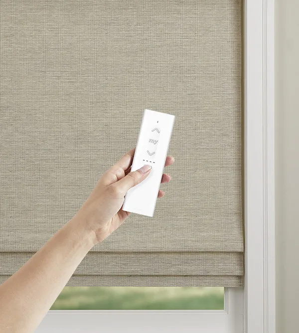 Hand holding the Somfy Ysia 5 channel remote control in front of a Natural Woven shade.