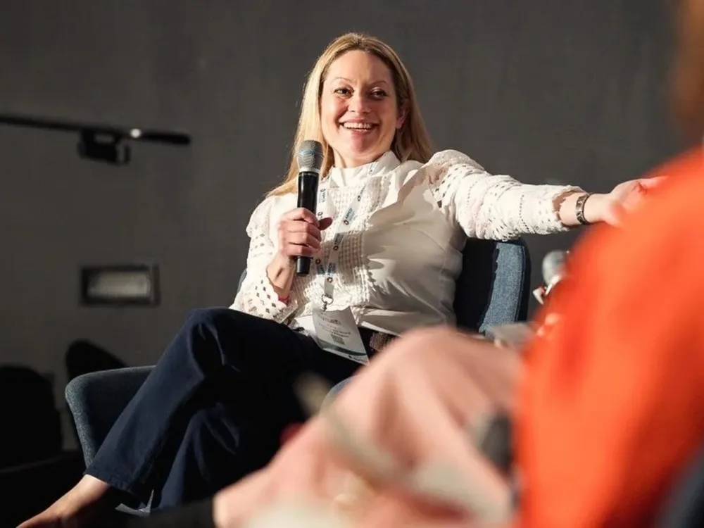 A woman sitting on a chair, holding a microphone and extending her hand towards someone off-camera. She is wearing a white blouse and dark trousers.