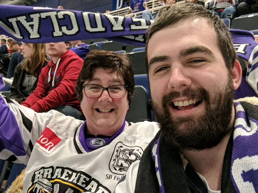 Craig and his mother wearing hockey jerseys raise their scarves, celebrating their team with excitement.