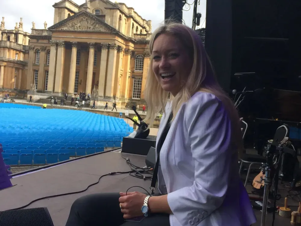 A woman with long blonde hair wearing a white blazer sits on a stage, smiling. In the background, a large historic building is visible, along with a blue stage setup.