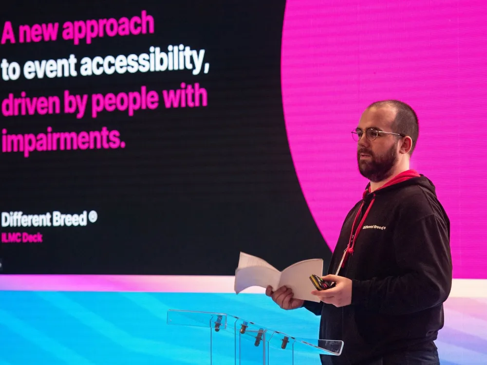 A man in glasses stands at a podium, presenting to an audience at a professional conference.