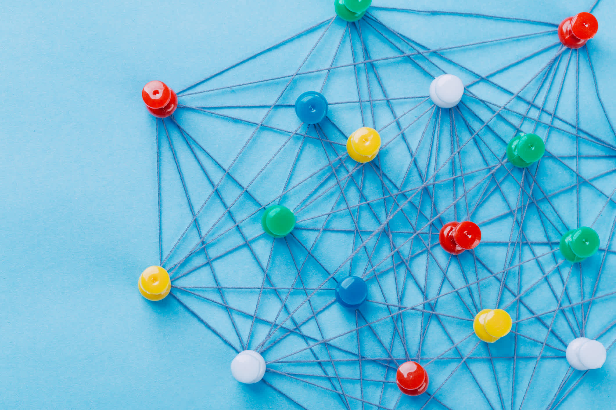 A small network of pins (Thumbtack)and string, An arrangement of colorful pins linked together with string on a pale blue background, suggests a network of connections. | Watermark