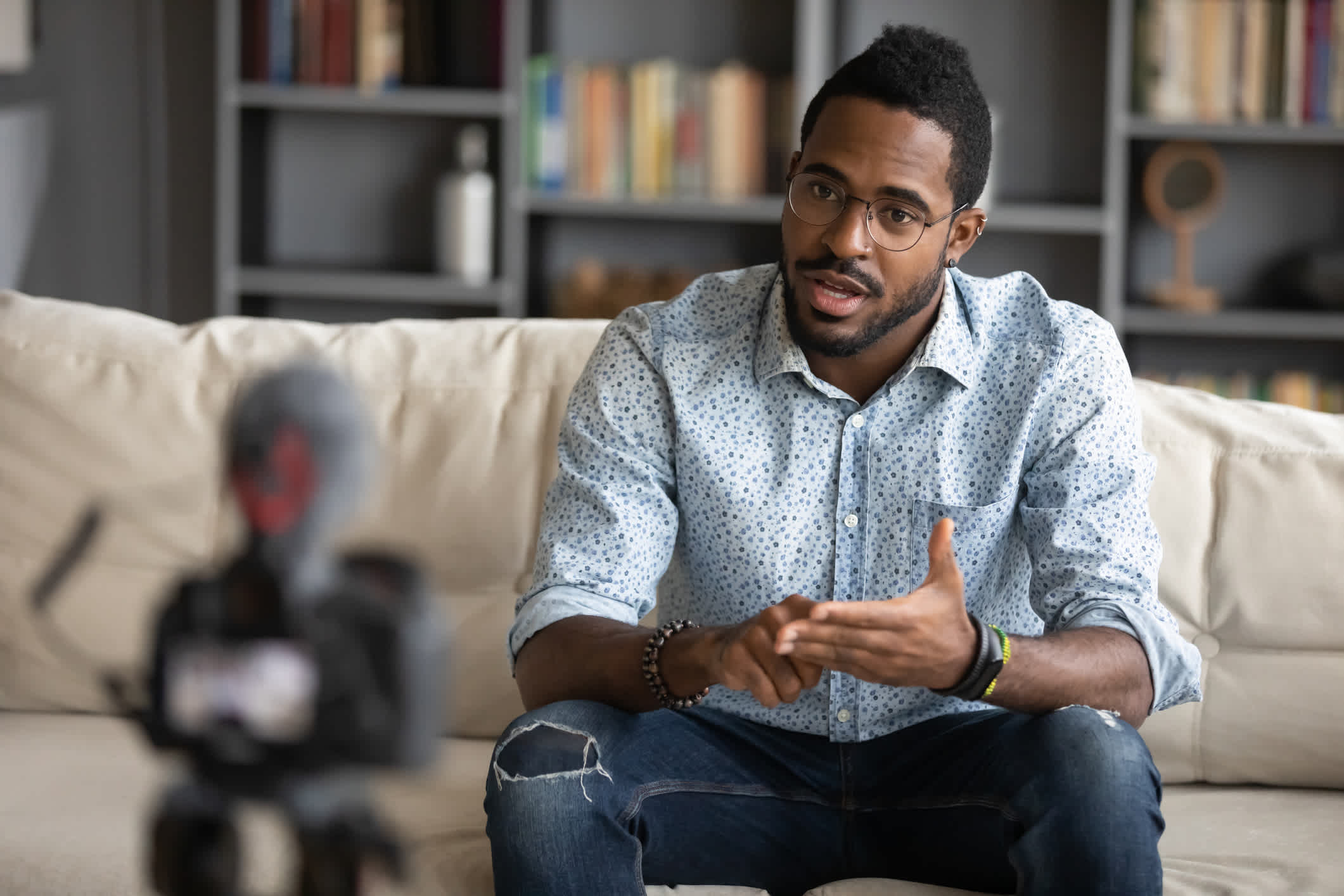 African American man sitting on couch filming online training webinar using camcorder dslr modern professional device camera. Value proposition. | Watermark