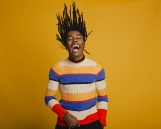 Waist-up portrait of a young man flipping his hair in front of a yellow background. He is wearing a striped sweater.