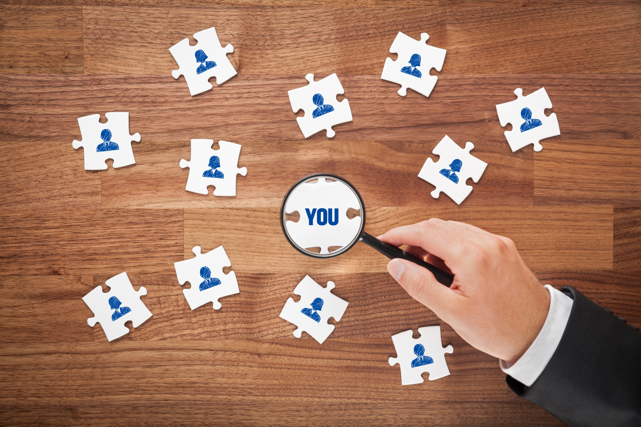 Personalization concept: A hand holding a magnifying glass focuses on a puzzle piece with the word 'you,' symbolizing individuality among other puzzle pieces with illustrated silhouettes of people's portraits.| Watermark