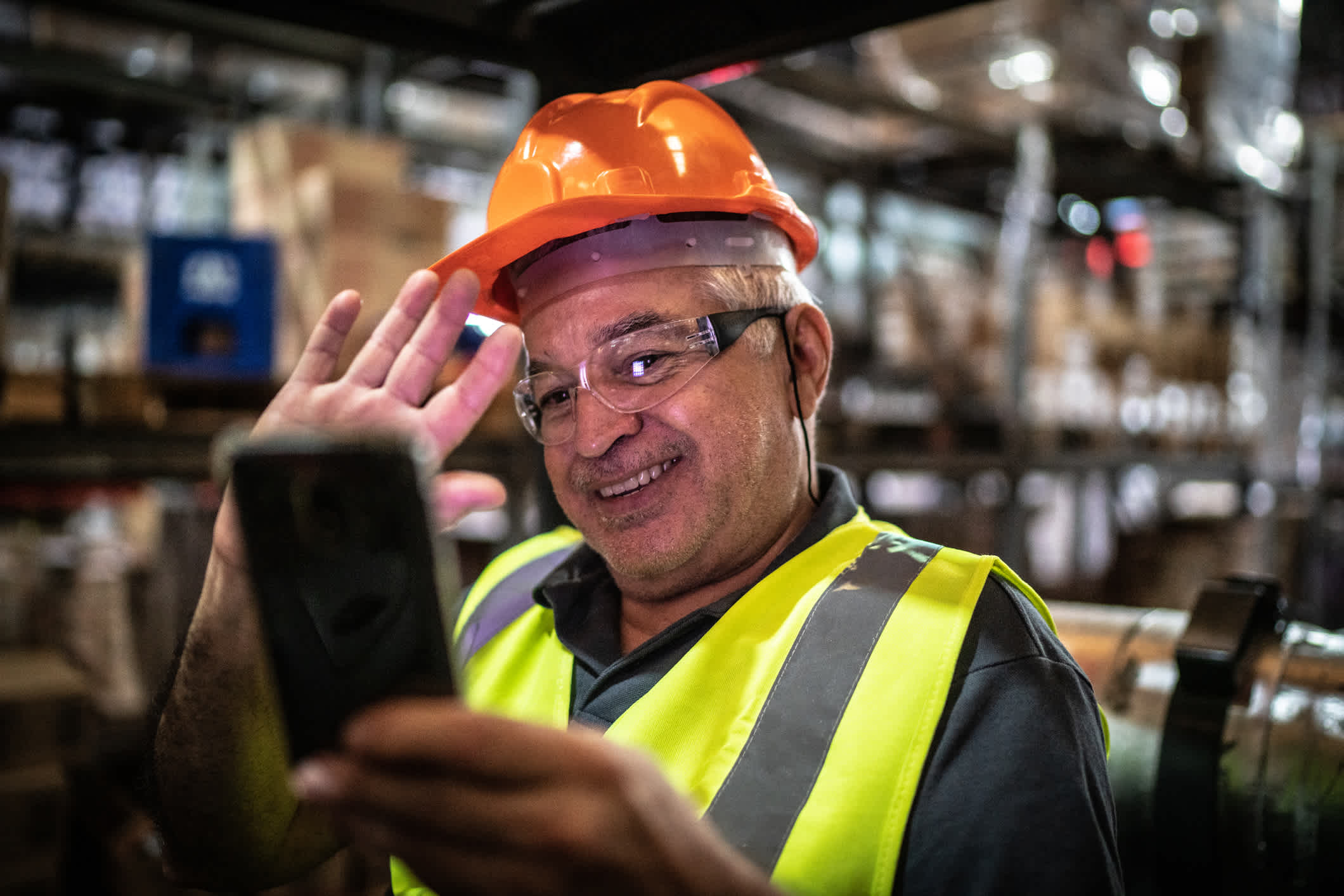 Warehouse worker with a hardhat on holding a phone to record a video in the warehouse. Use-case for video marketing in the manufacturing sector. | Watermark