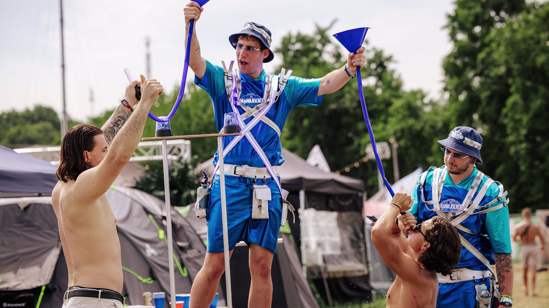 Zwei Festivalbesucher trinken aus blauen Trichtern, die von einem als ‚Brandlöscher‘ gekleideten Teammitglied auf einem Podest gehalten werden; im Hintergrund Zelte und Sommerstimmung.