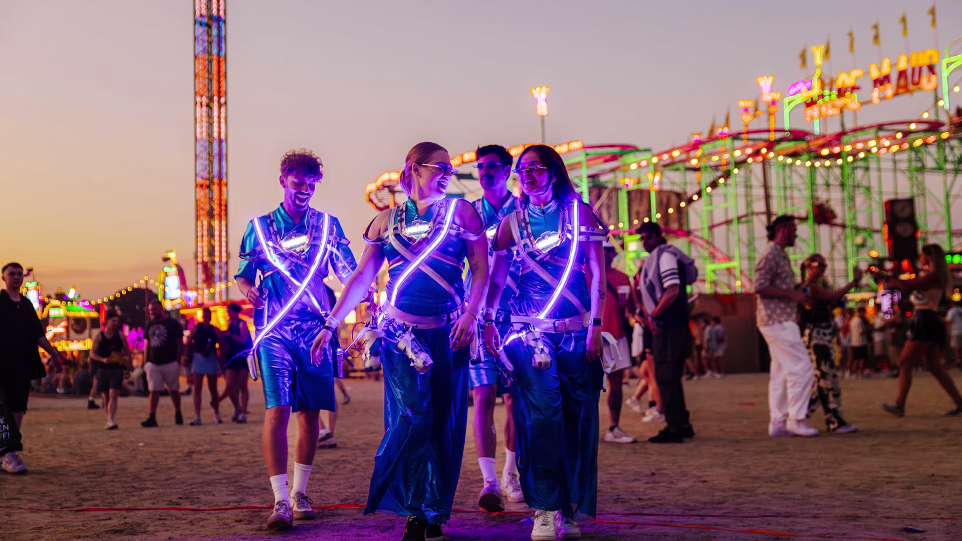 Vier Brandlöscher-Promoter in leuchtenden Outfits laufen lachend über ein bunt erleuchtetes Festivalgelände bei Sonnenuntergang – im Hintergrund Achterbahn und Besucher.