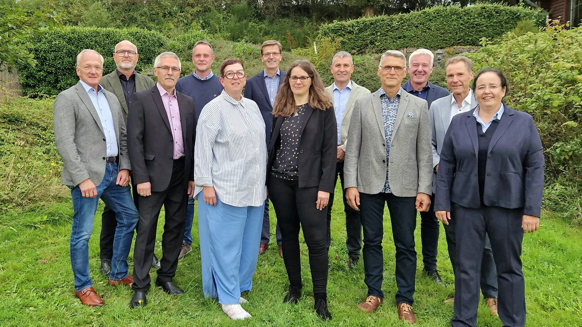 Gruppenfoto der Gerolsteiner-Jubilare 2024: 13 Männer und Frauen unterschiedlichen Alters posieren lächelnd im Grünen, festlich gekleidet anlässlich ihres Dienstjubiläums.