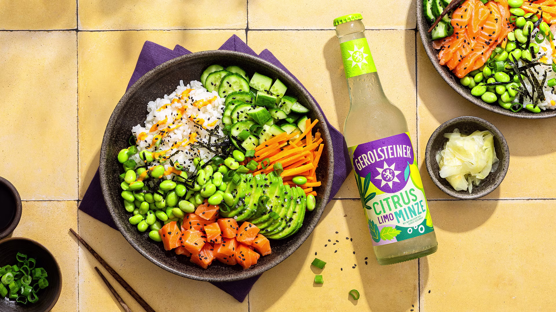 Draufsicht auf eine farbenfrohe Sushi-Bowl mit Reis, Edamame, Lachs, Avocado, Gurke und Karotten auf einem hellen Fliesentisch. Daneben steht eine Flasche Gerolsteiner Citrus Limo Minze.