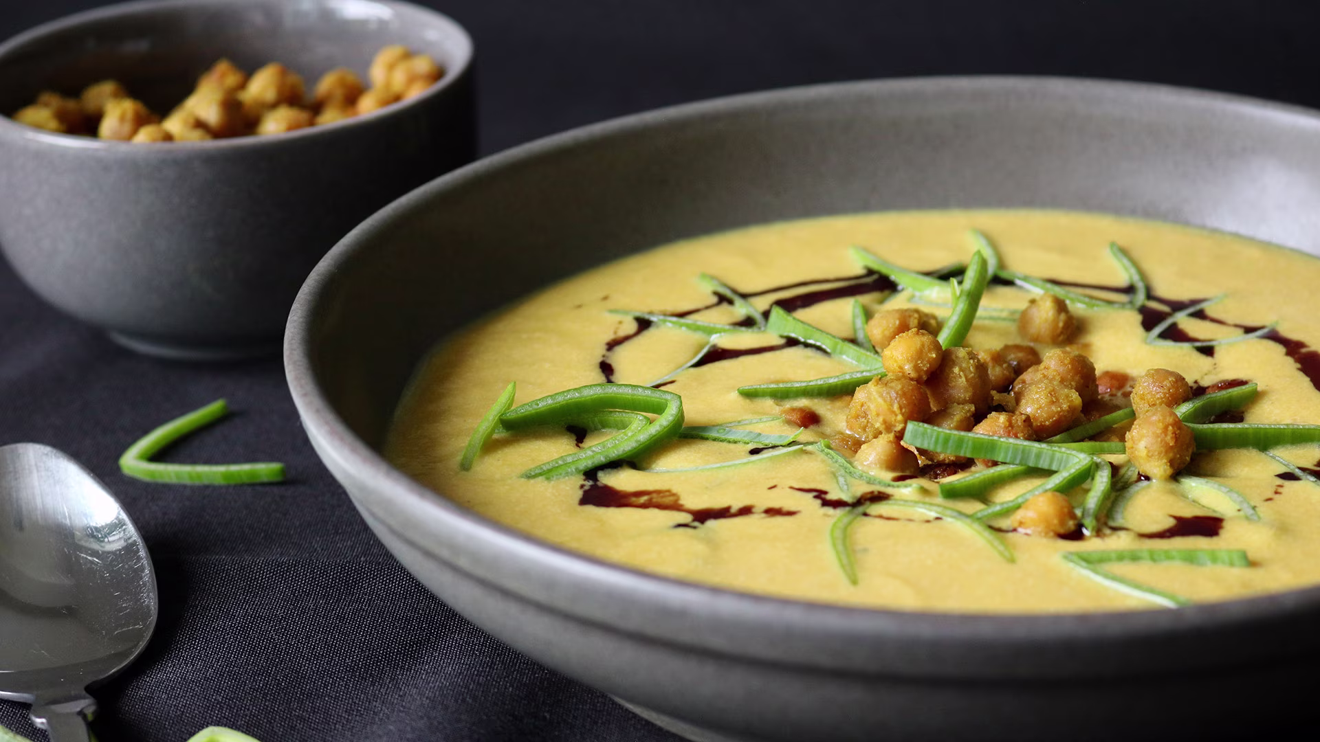 Cremige Kürbissuppe in einer dunklen Schale, garniert mit knusprigen Kichererbsen, feinen Lauchstreifen und Balsamico-Creme, daneben eine Schüssel mit gerösteten Kichererbsen auf schwarzer Tischdecke.