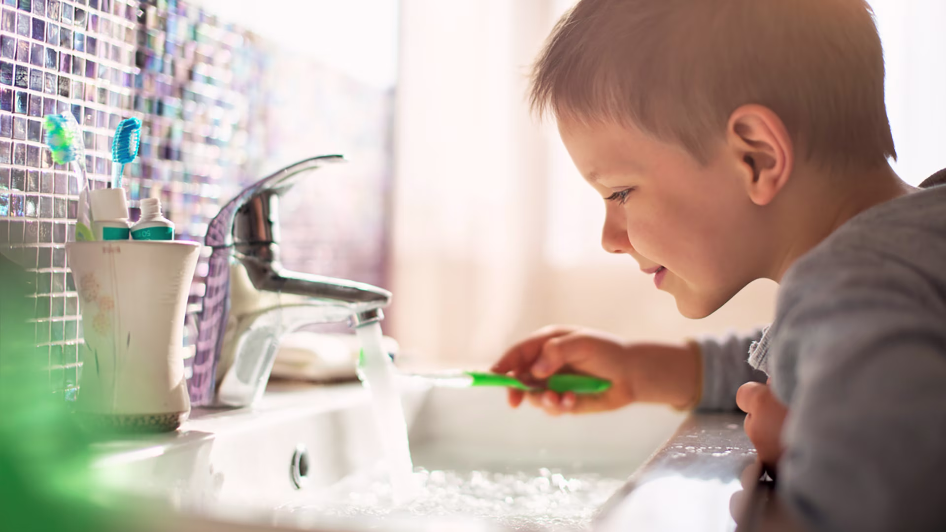 Kleiner Junge will sich die Zähne putzen und hält seine Zahnbürste unter den fließenden Wasserhahn im Badezimmer.