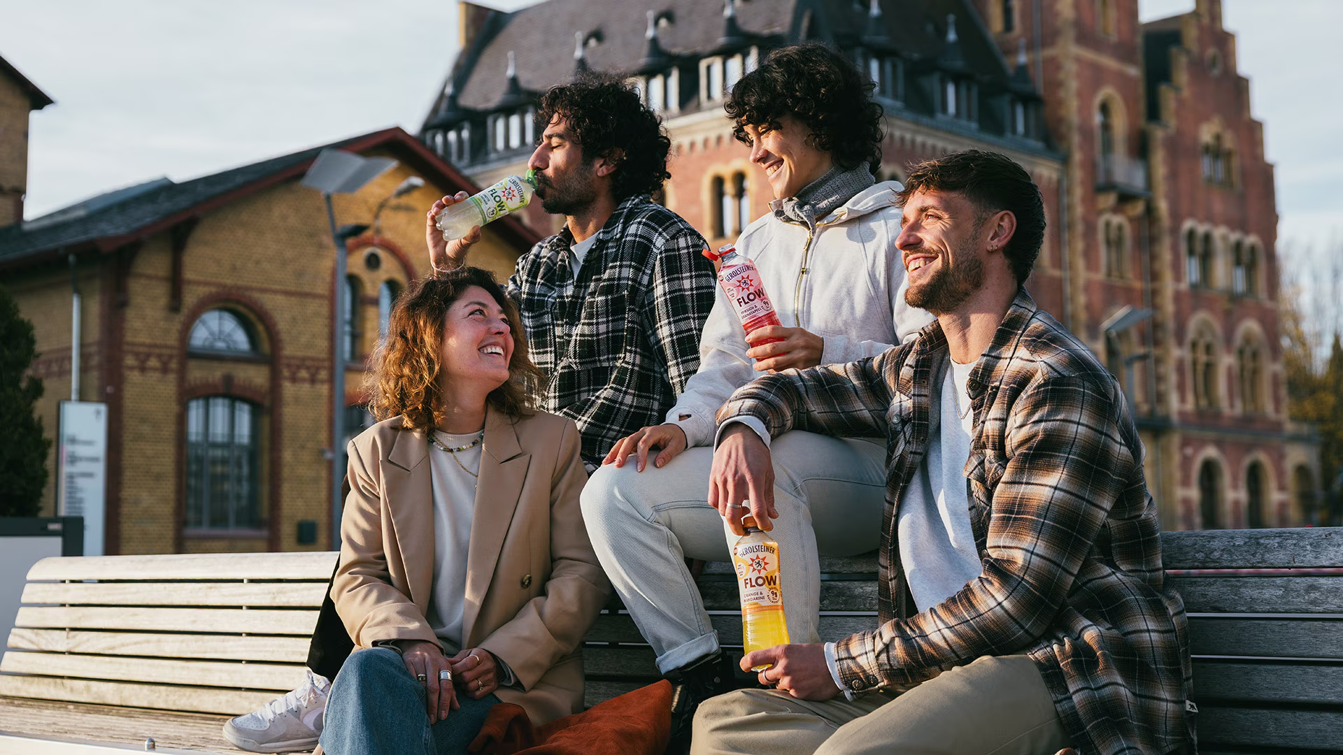 Vier junge Erwachsene sitzen entspannt auf einer Bank vor einem historischen Gebäude, lachen und trinken verschiedene Sorten Gerolsteiner Flow. Die warme Sonne sorgt für eine lockere, fröhliche Stimmung.