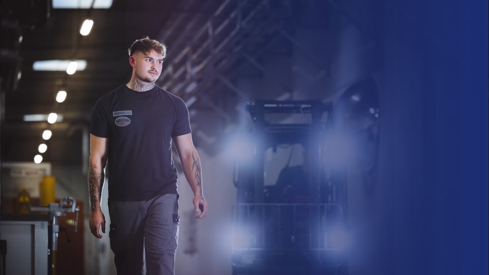 Ein junger Mann in Arbeitskleidung läuft in einer Werkshalle entlang. Im Hintergrund sieht man einen Gabelstapler.