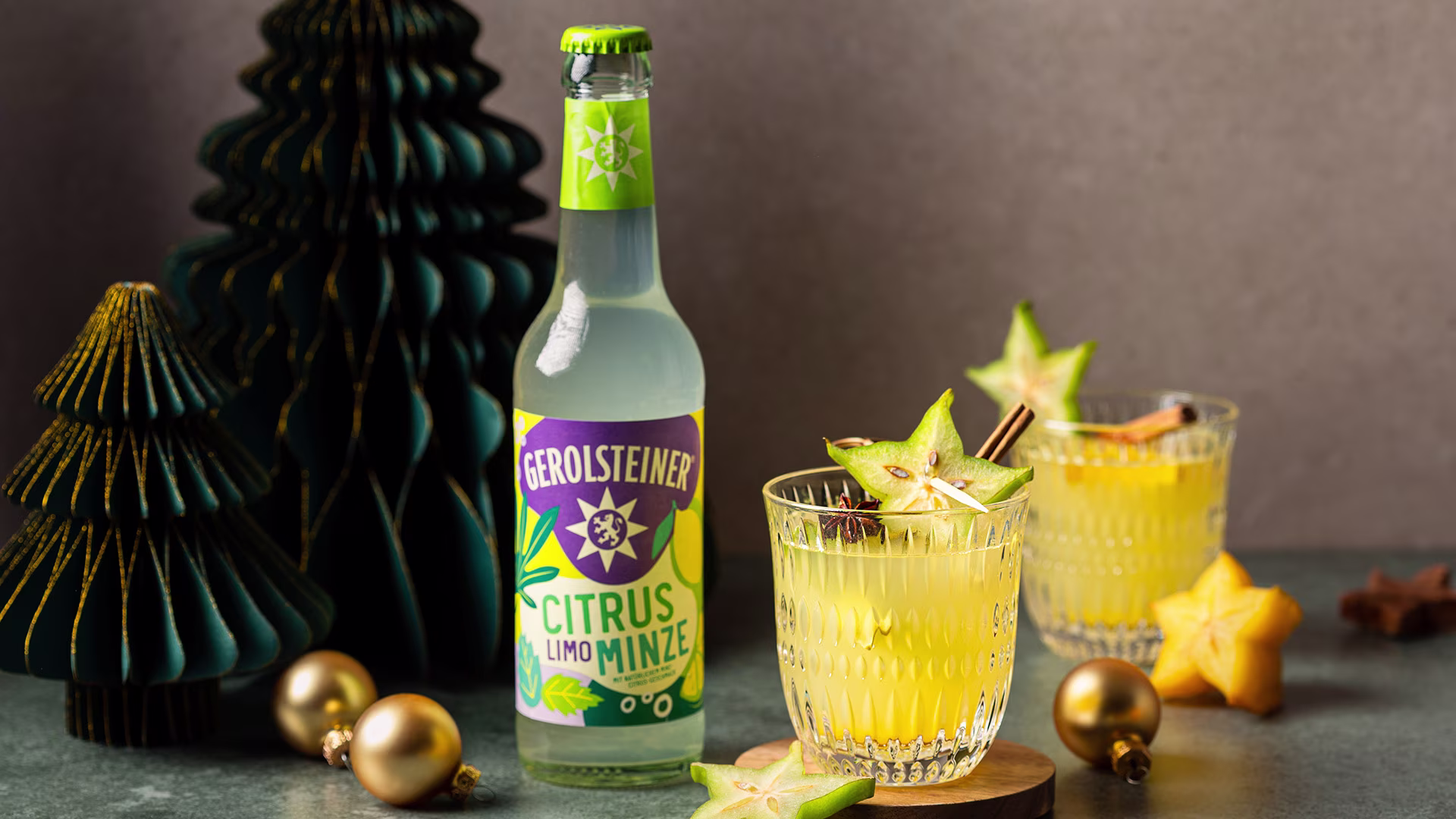 Eine Flasche Gerolsteiner Citrus Limo Minze steht neben zwei Gläsern mit gelbem Getränk, garniert mit Sternfrucht und Zimtstange. Goldene Christbaumkugeln und grüne Deko-Tannen sorgen für eine festliche, winterliche Atmosphäre.
