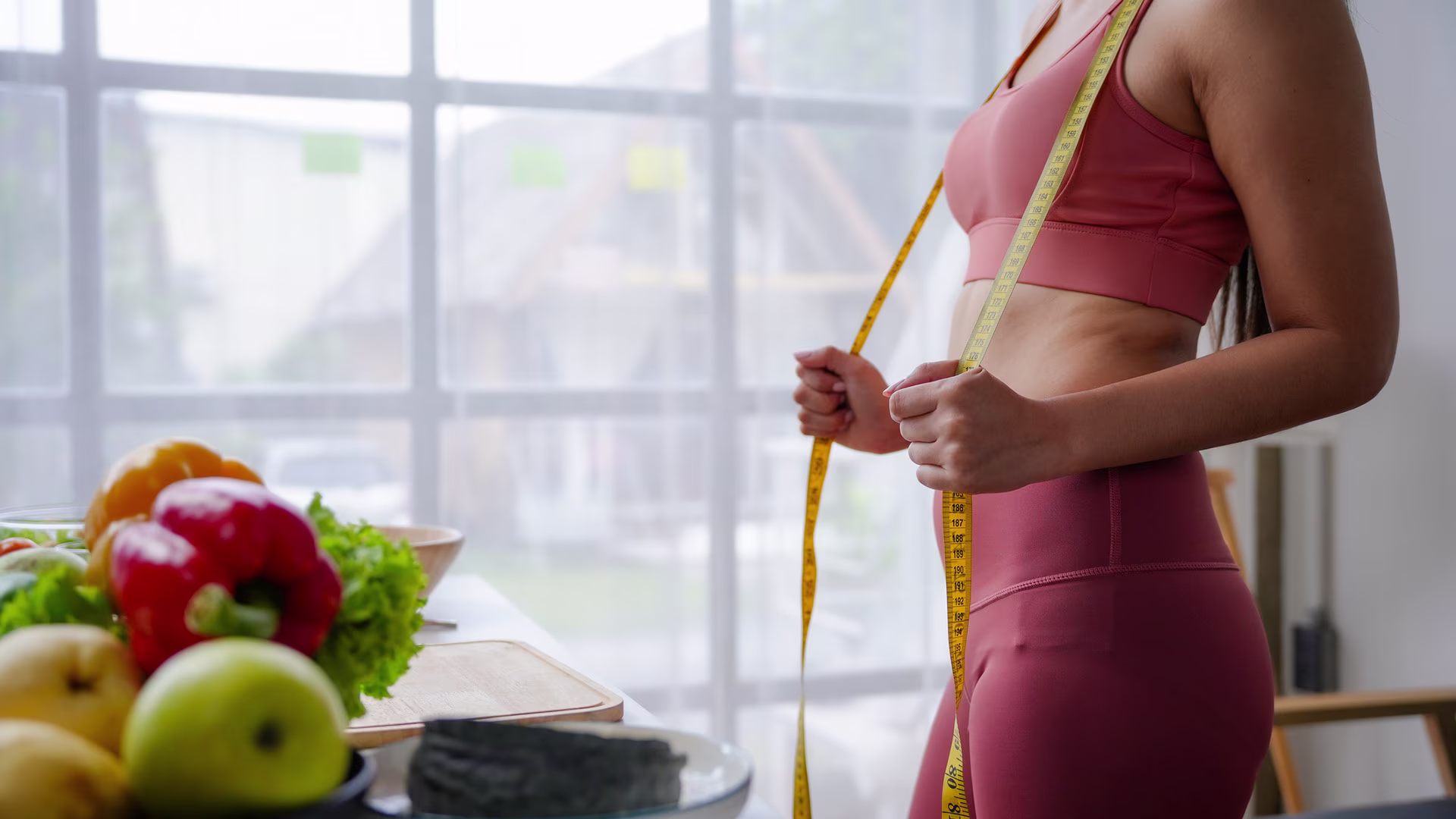Frau misst zu Hause ihre Taille mit einem Maßband neben frischem Obst, Symbol für Erfolgsmessung und Anpassung des Ernährungsplans bei einer Ernährungsumstellung, um Fortschritte wie Gewichtsverlust und gesundheitliche Verbesserungen zu verfolgen.
