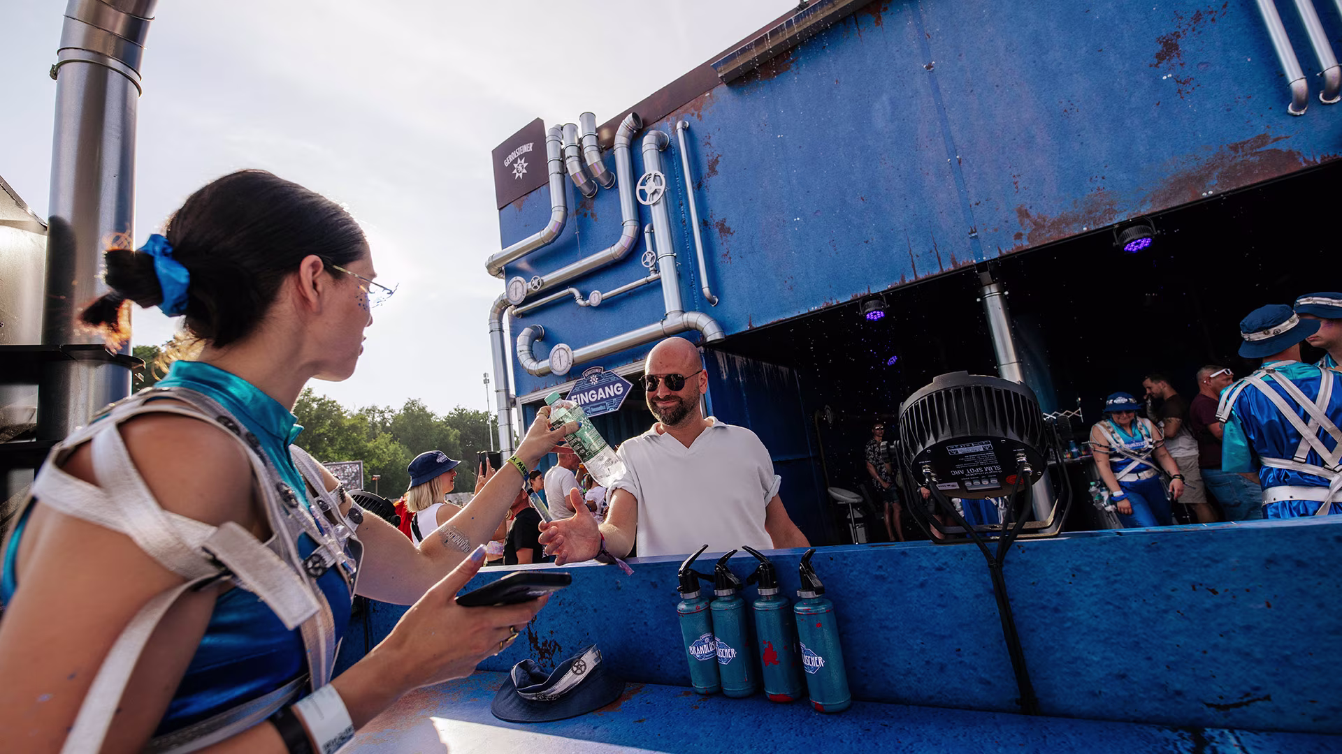 Festivalbesucher erhält eine Flasche Gerolsteiner von einer als Brandlöscher verkleideten Promoterin am blauen Verkaufsstand mit futuristischen Rohrleitungen.