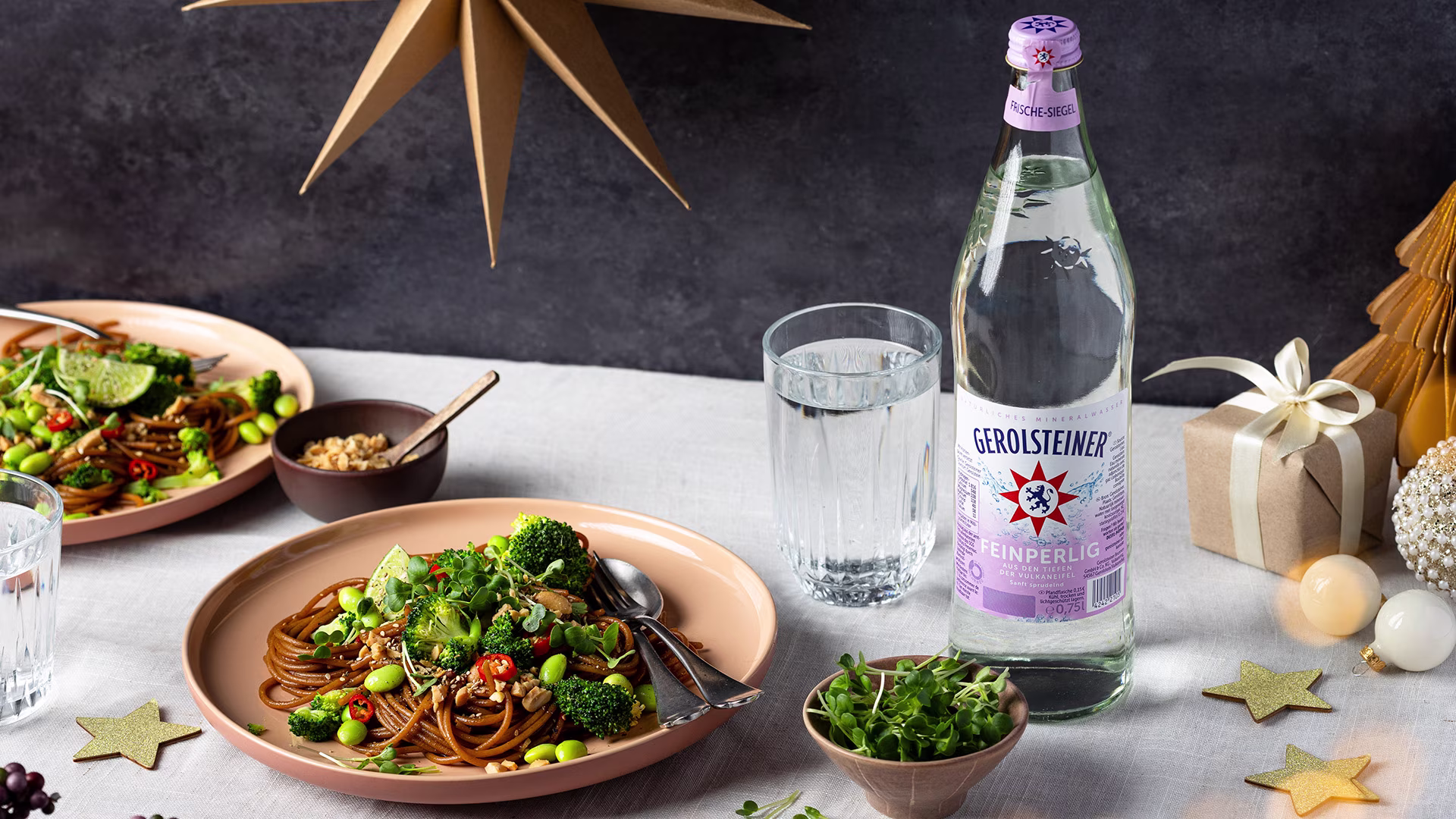 Ein festlich gedeckter Tisch mit Tellern voll Asia-Vollkornnudeln, Brokkoli und Edamame. Daneben steht eine Flasche Gerolsteiner Feinperlig und ein Glas Wasser. Dekoration mit Sternen, Geschenk und Lichtern sorgt für eine gemütliche, festliche Atmosphäre.