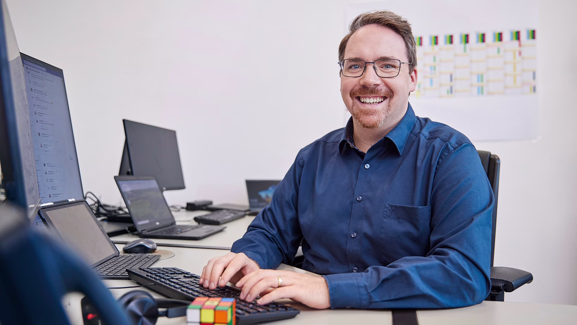 Lächelnder Mitarbeiter im IT-Bereich sitzt an einem Arbeitsplatz mit mehreren Monitoren und Laptops, tippt auf einer Tastatur; im Vordergrund ein Rubik’s Cube und Headset.
