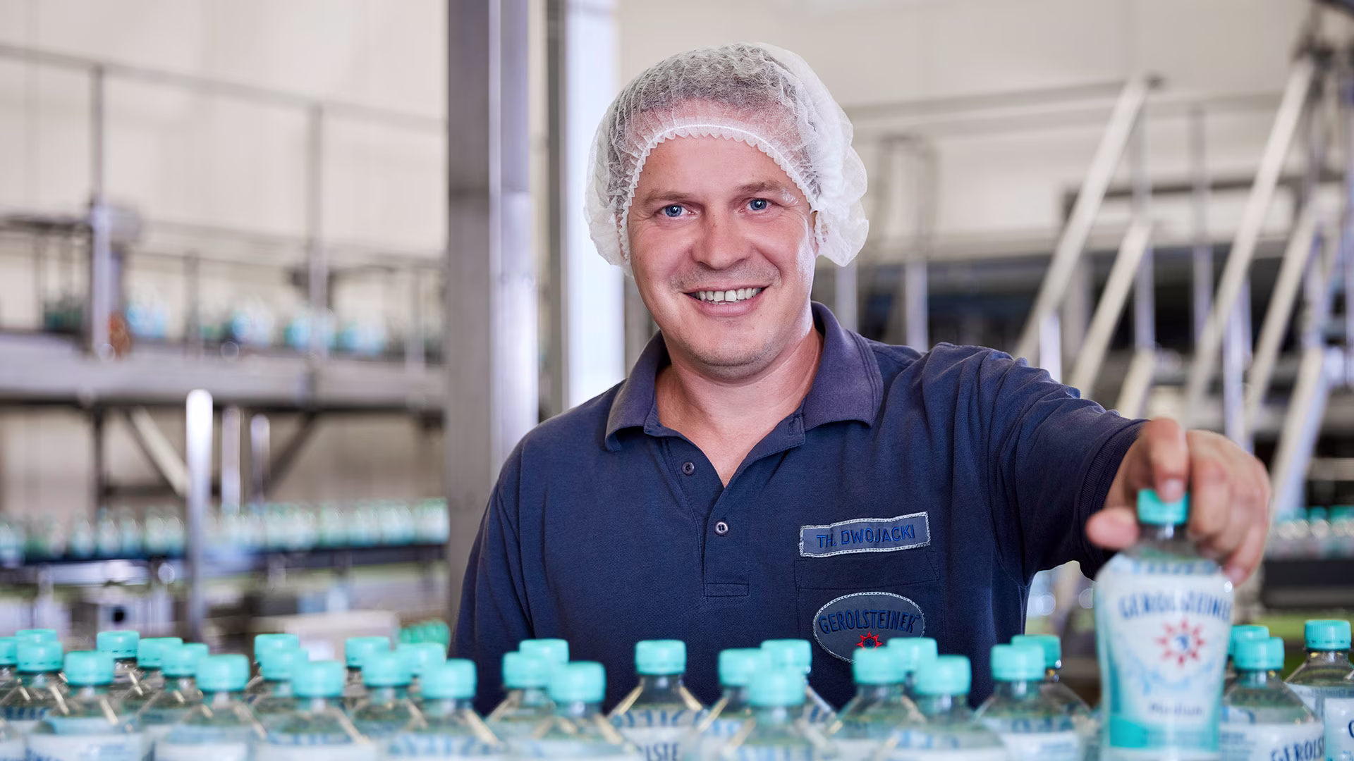 Mitarbeiter in Arbeitskleidung und Haarnetz steht lächelnd in einer Abfüllanlage vor mehreren Gerolsteiner-Flaschen und hält eine Flasche in der Hand.