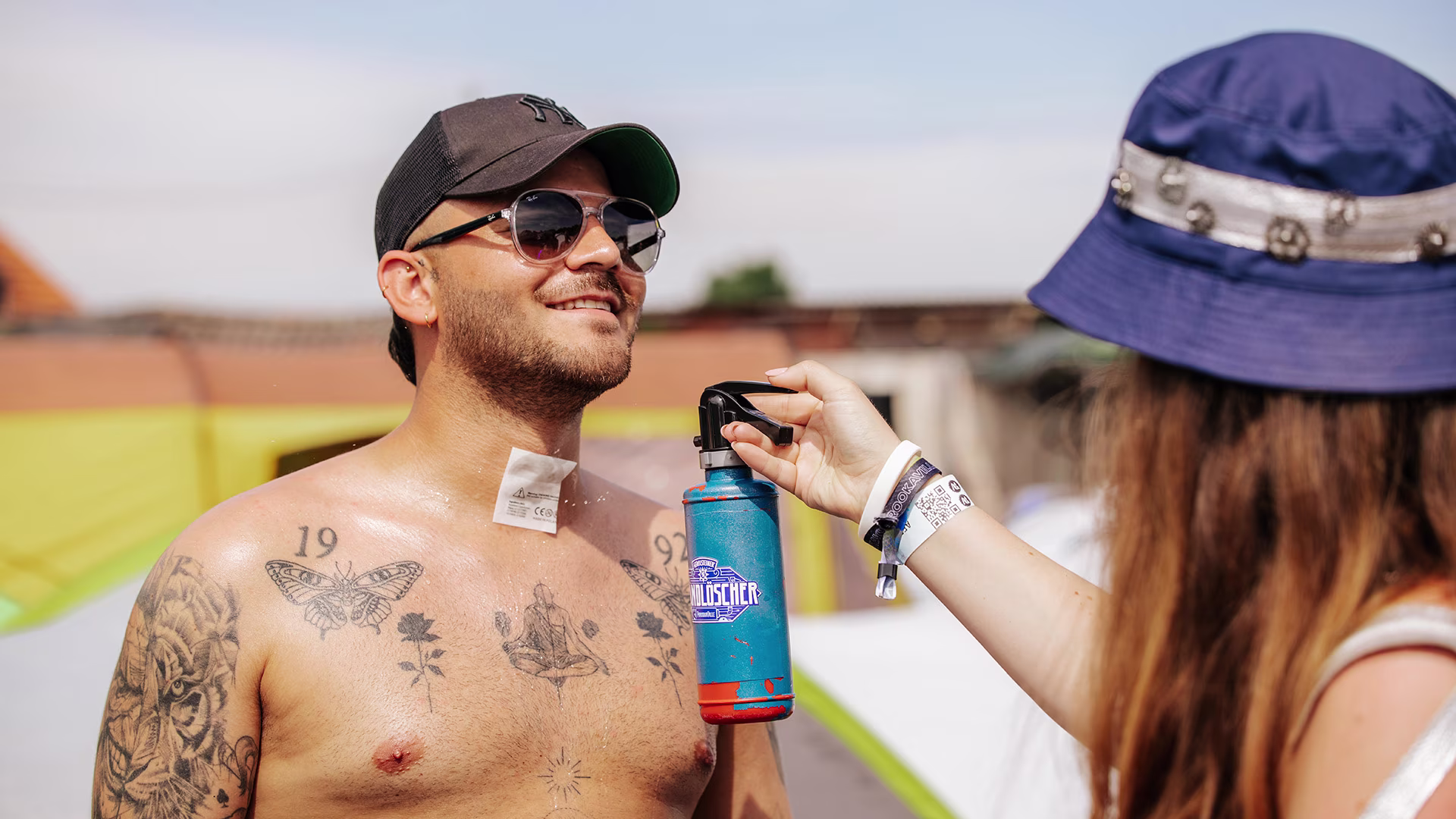 Festivalbesucher mit Sonnenbrille und Tattoos wird mit einer Brandlöscher-Sprühflasche von einer Person im Sonnenhut erfrischt – Sommerszene im Campingbereich.