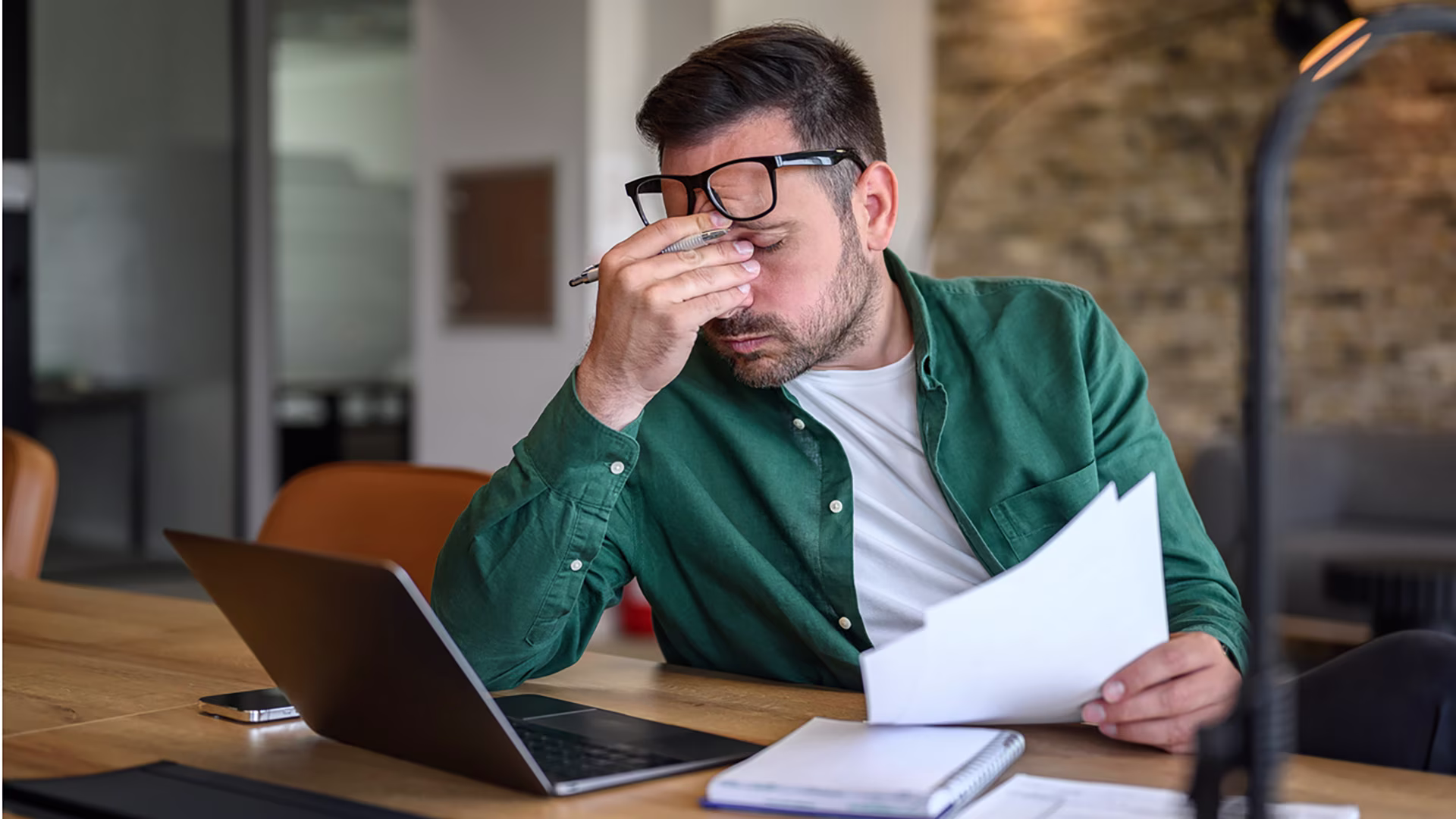 Ein Mann sitzt am Schreibtisch vor seinem Laptop und hält ein Dokument in der Hand. Er reibt sich gestresst die Augen.