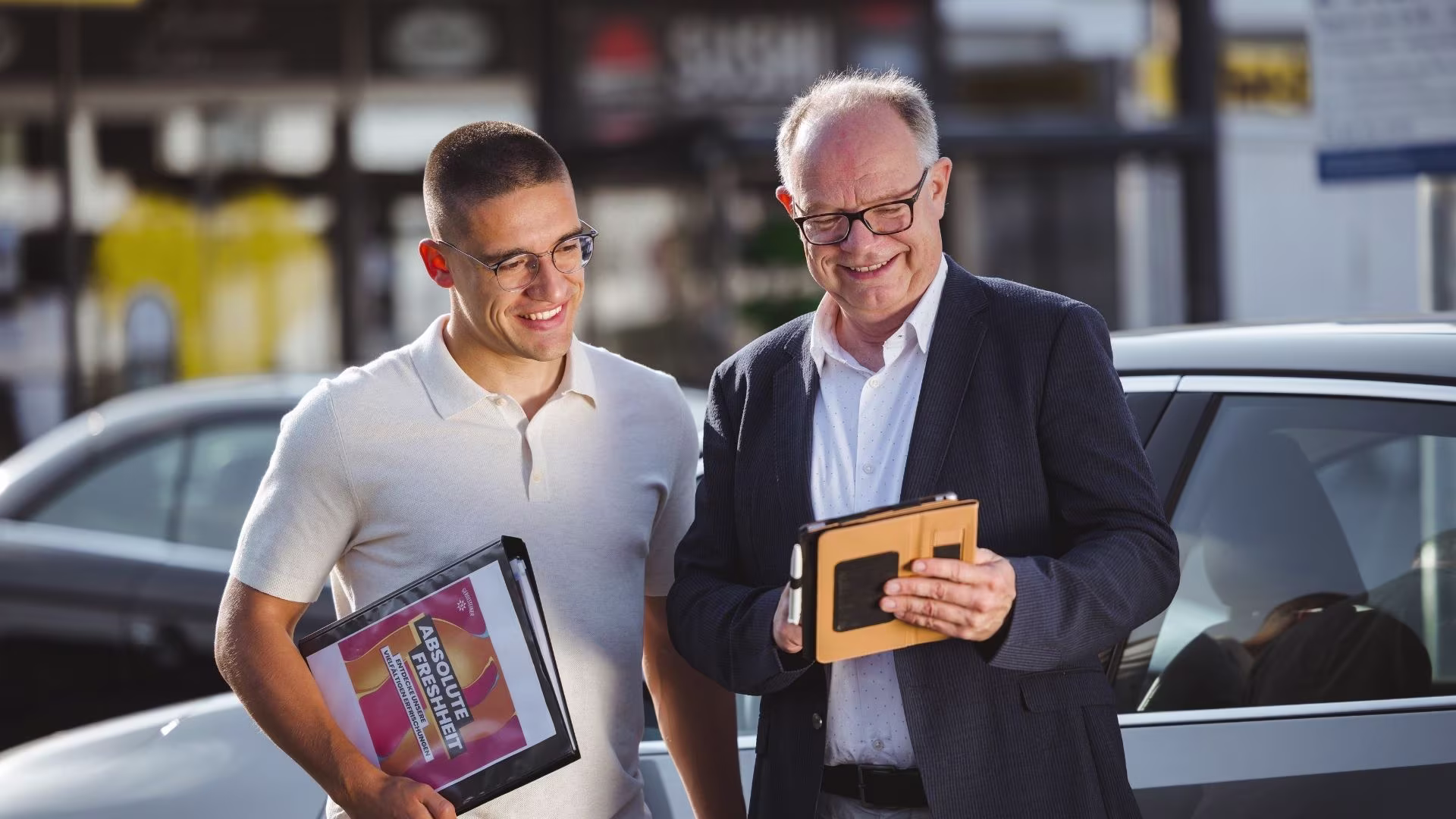 Zwei Mitarbeitende mit Brille führen ein Gespräch und schauen dabei auf ein iPad – im Hintergrund ist ein Fahrzeug zu sehen.