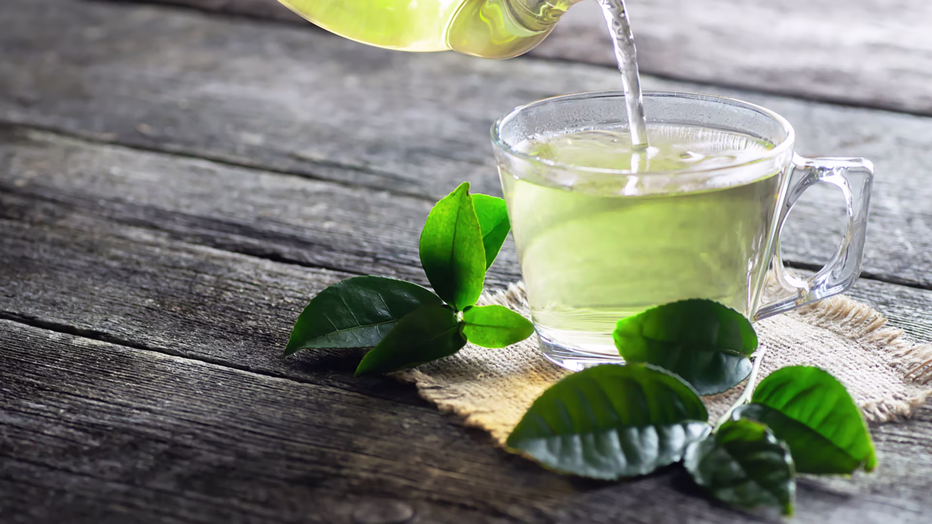 Grüner Tee wird aus Glaskanne in auf Holztisch stehende durchsichtige Tasse gegossen