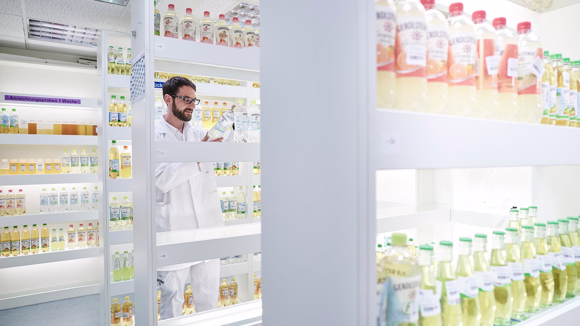 Mitarbeiter in weißem Laborkittel prüft eine Getränkeflasche in einem hell ausgeleuchteten Raum mit Regalen voller Produktproben zur Qualitätssicherung.