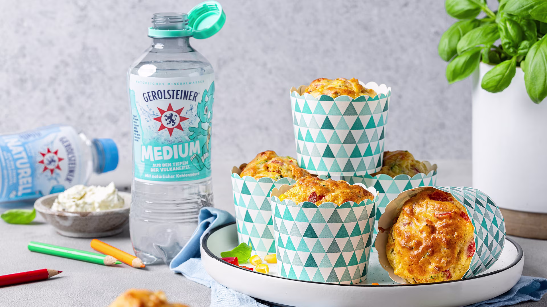 Gerolsteiner Medium PET-Flasche neben frisch gebackenen Gemüse-Pizzamuffins in Papierförmchen auf einem Teller. Im Hintergrund stehen Basilikum, Frischkäse und eine weitere Wasserflasche – eine Idee für die Brotdose.