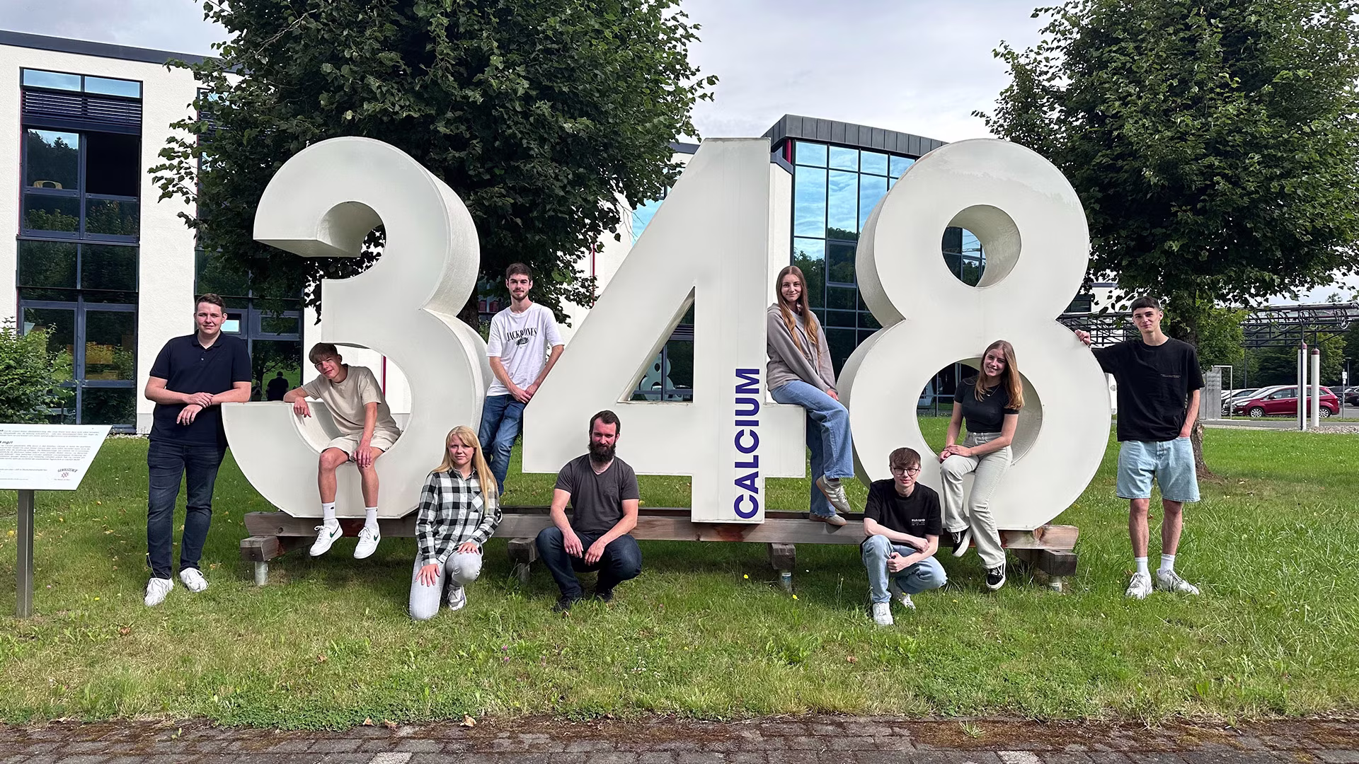 Gruppenfoto von neun jungen Auszubildenden vor der großen weißen Zifferninstallation ‚348 CALCIUM‘ auf dem Firmengelände von Gerolsteiner – aufgenommen zum Start ihrer Ausbildung 2024.
