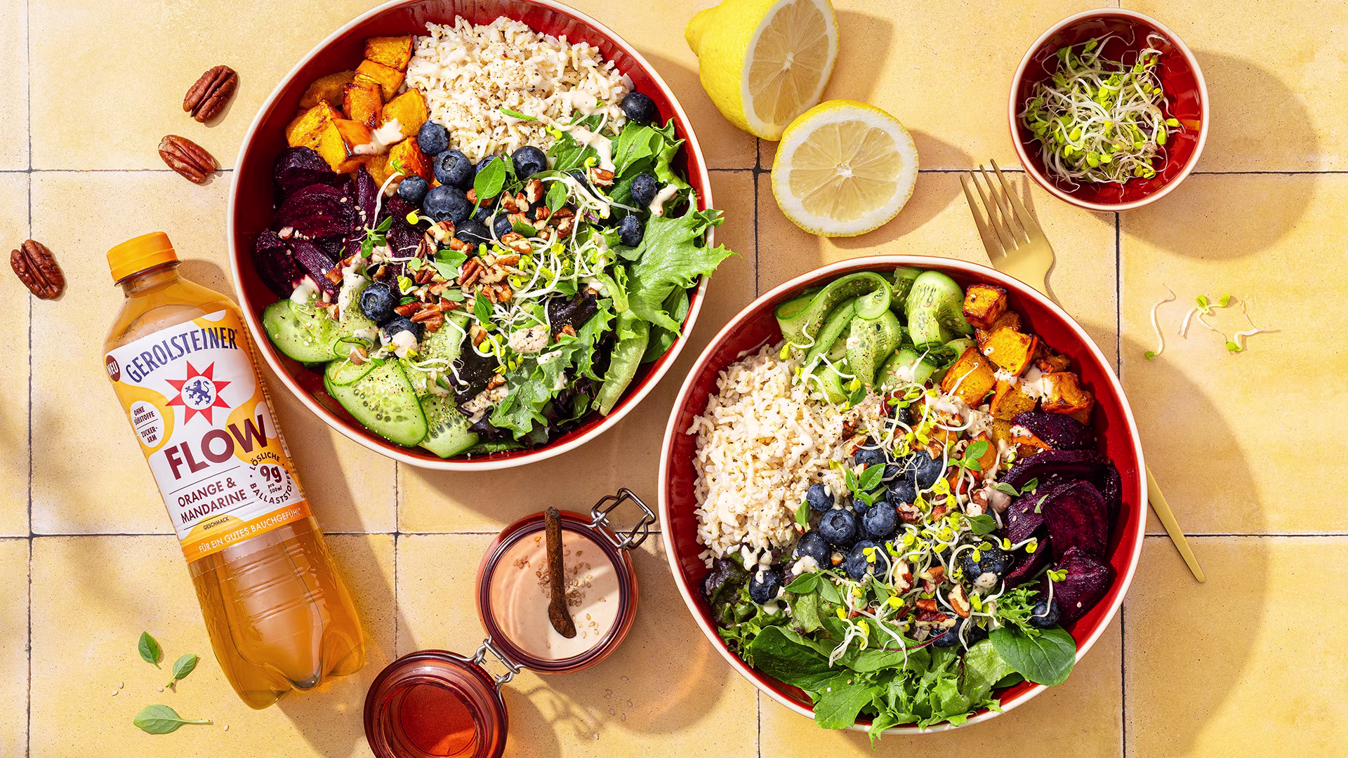 wei herbstliche Bowls mit Reis, Kürbis, Roter Bete, Gurke, Salat, Heidelbeeren, Sprossen und Pekannüssen. Daneben steht eine Flasche Gerolsteiner Flow Orange & Mandarine.