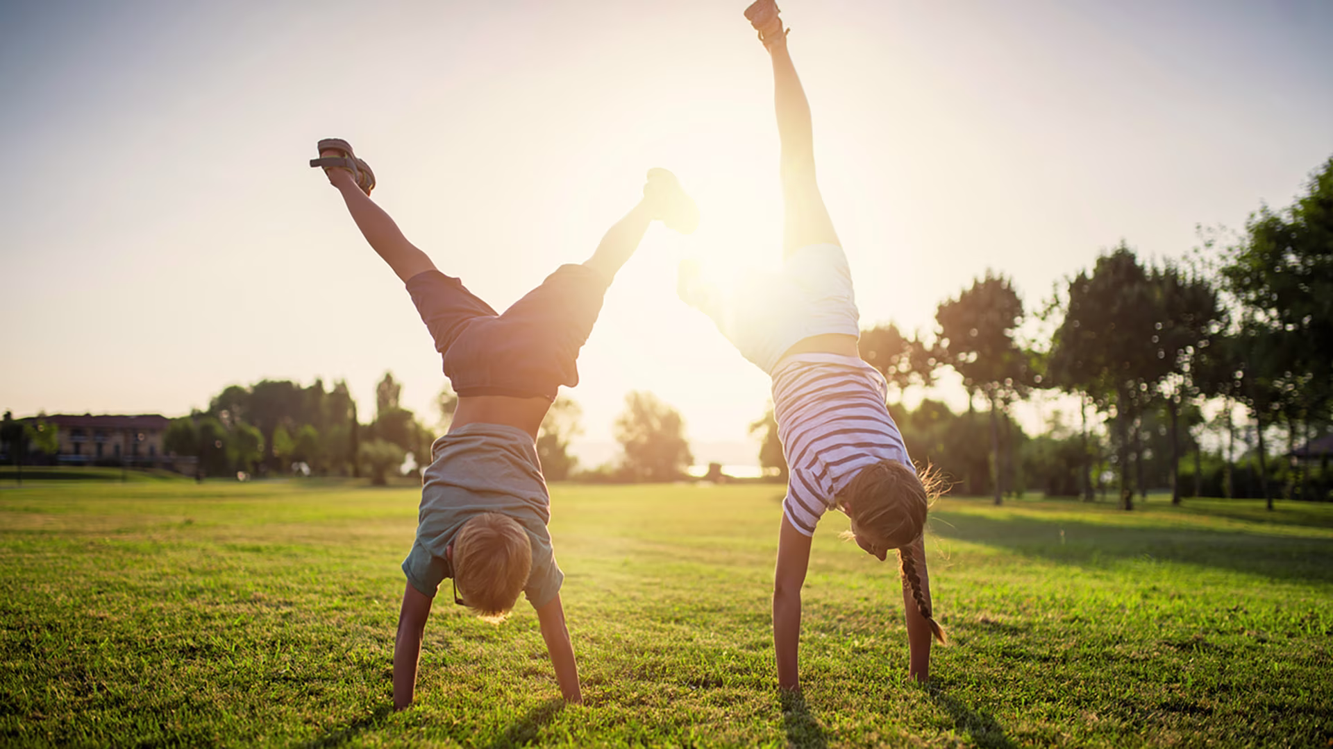 Zwei Kinder sind draußen in der Sonne auf einer Wiese und üben einen Handstand.