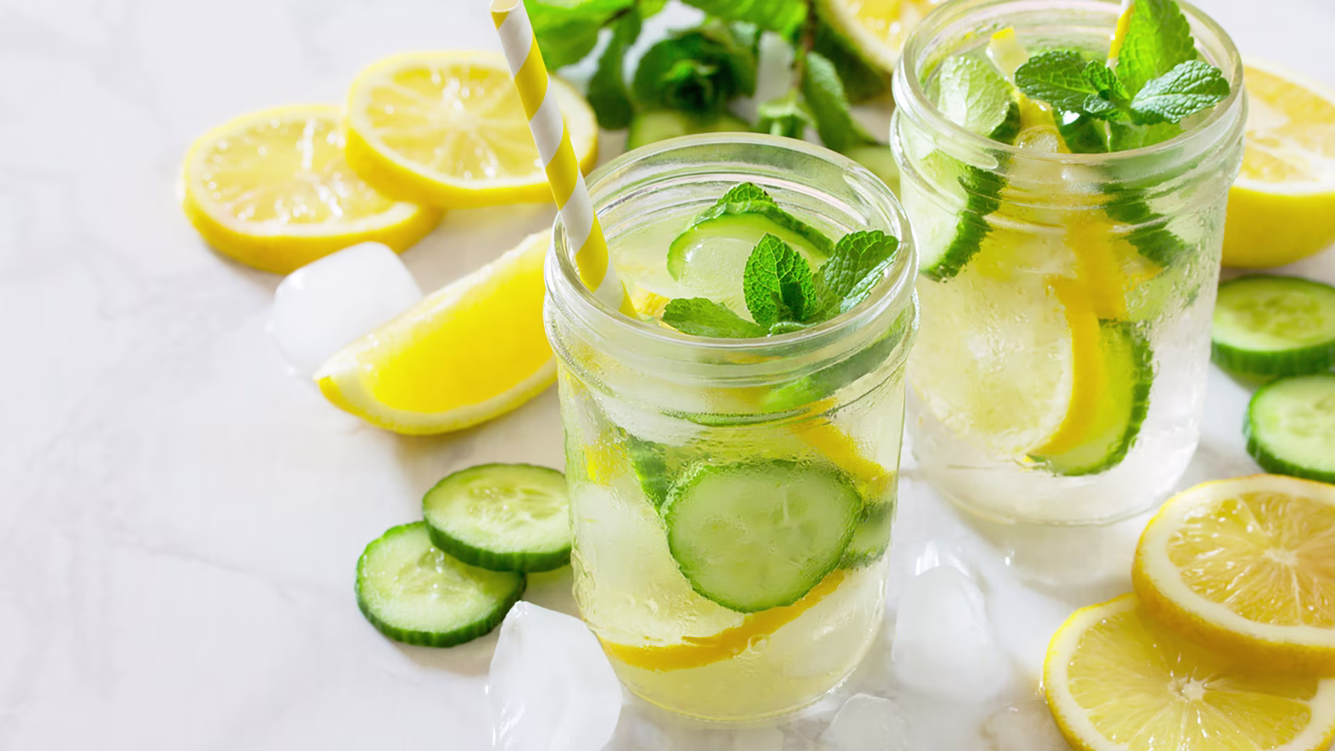 2 Gläser mit erfrischendem Gurkenwasser mit Gurken- und Zitronenscheiben und einem gelb-gestreiften Strohhalm.