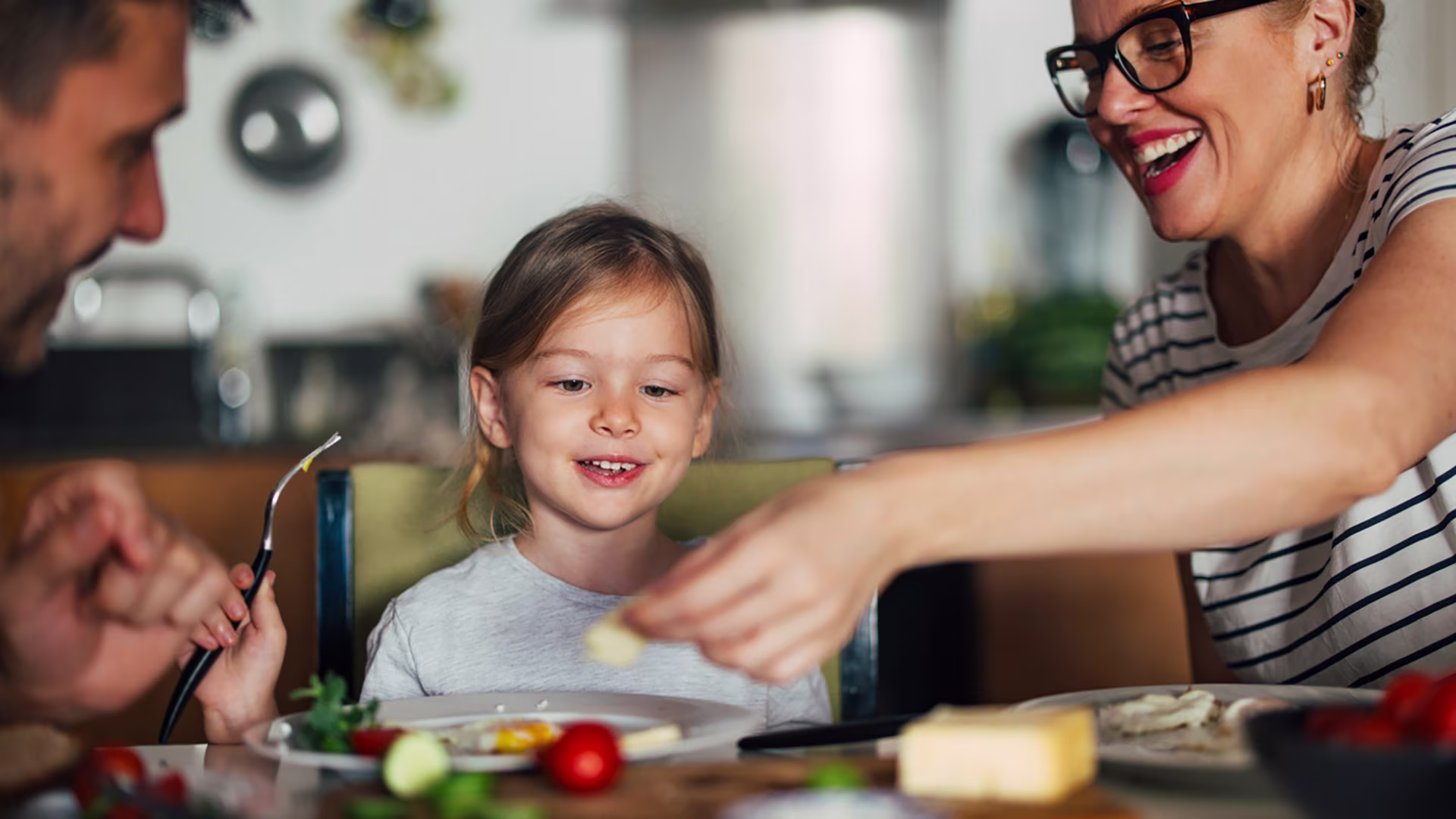 Glückliche Familie aus Mutter, Vater und Kind sitzen zuhause am Küchentisch und frühstücken. Das Kind ist im Fokus und blickt auf das Essen in der Hand der Mutter.