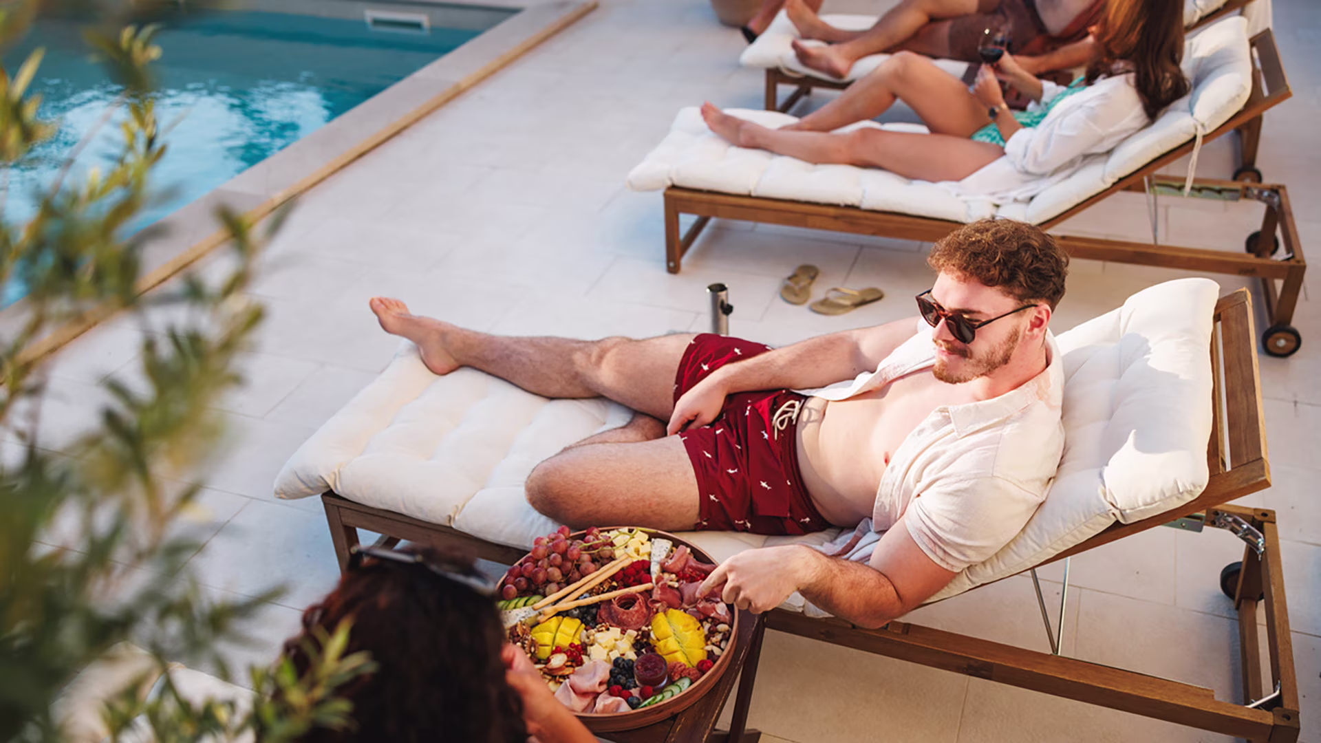 Freunde sitzen entspannt auf Liegen am Pool und trinken oder essen von einem gesunden Charcuterie Board.