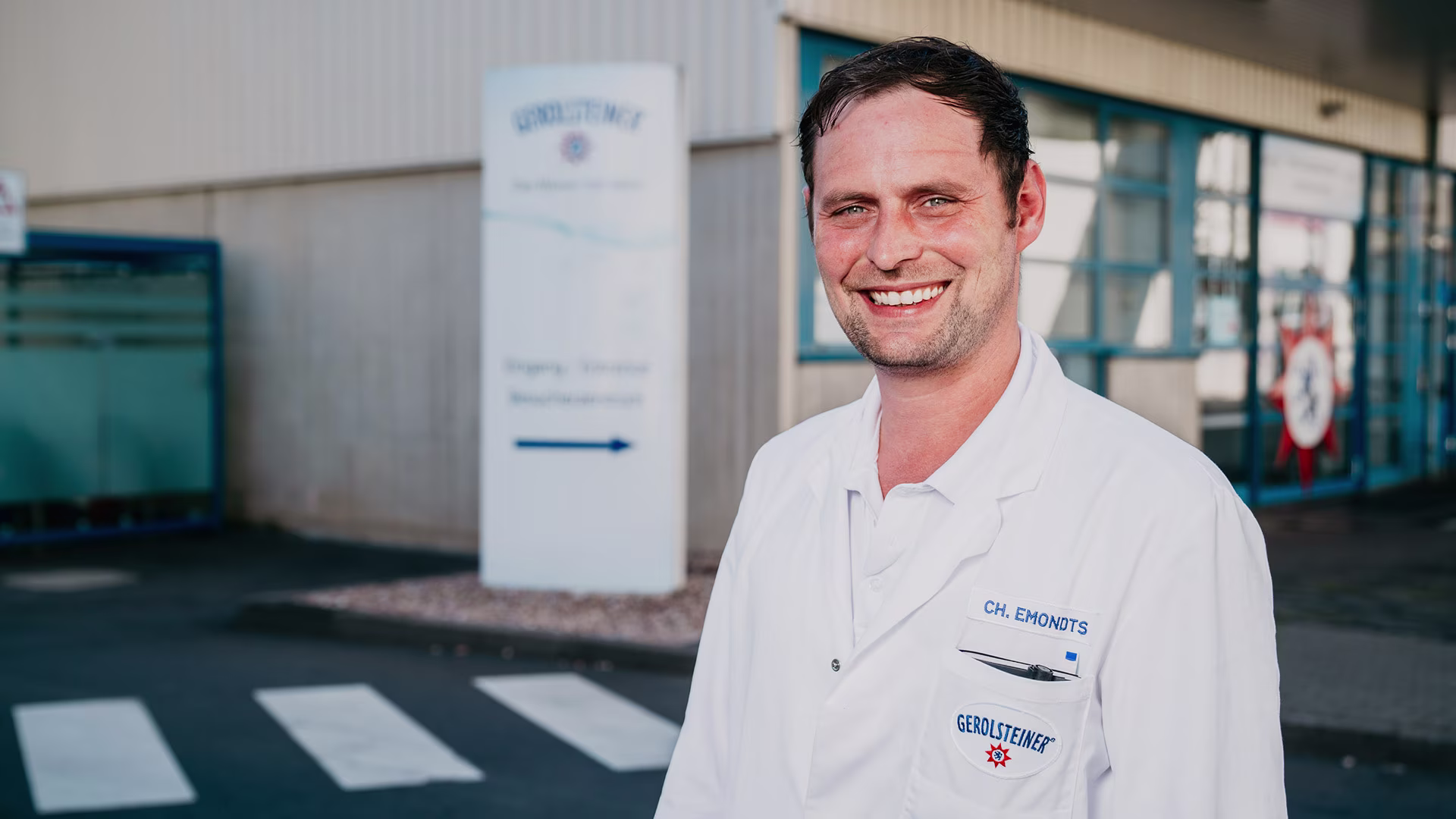Lächelnder Mitarbeiter in weißem Laborkittel mit Gerolsteiner-Logo steht vor dem Werksgebäude am Eingang, Zebrastreifen und Beschilderung im Hintergrund.