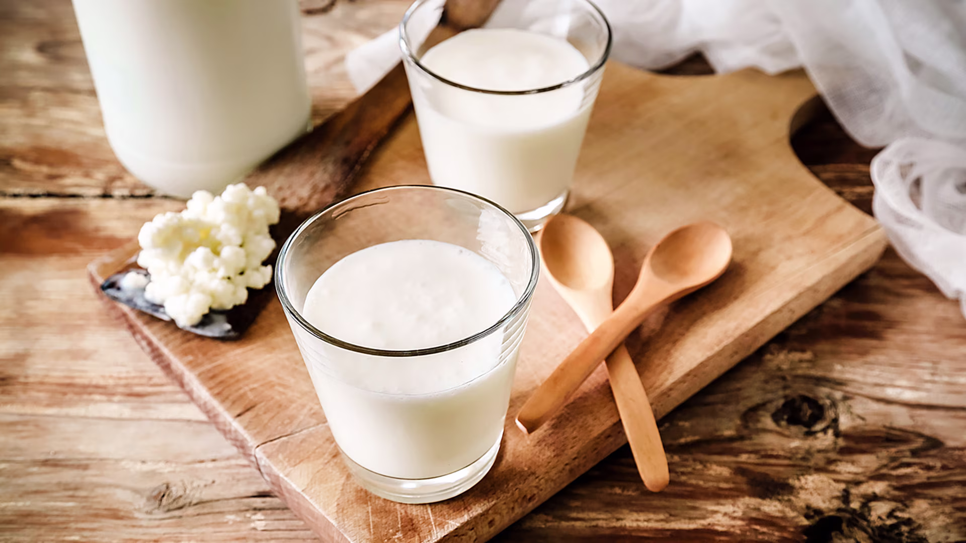 Zwei Gläser mit Kefir, Kefirknollen und kleine Holzlöffel liegen auf einem Holzbrett.