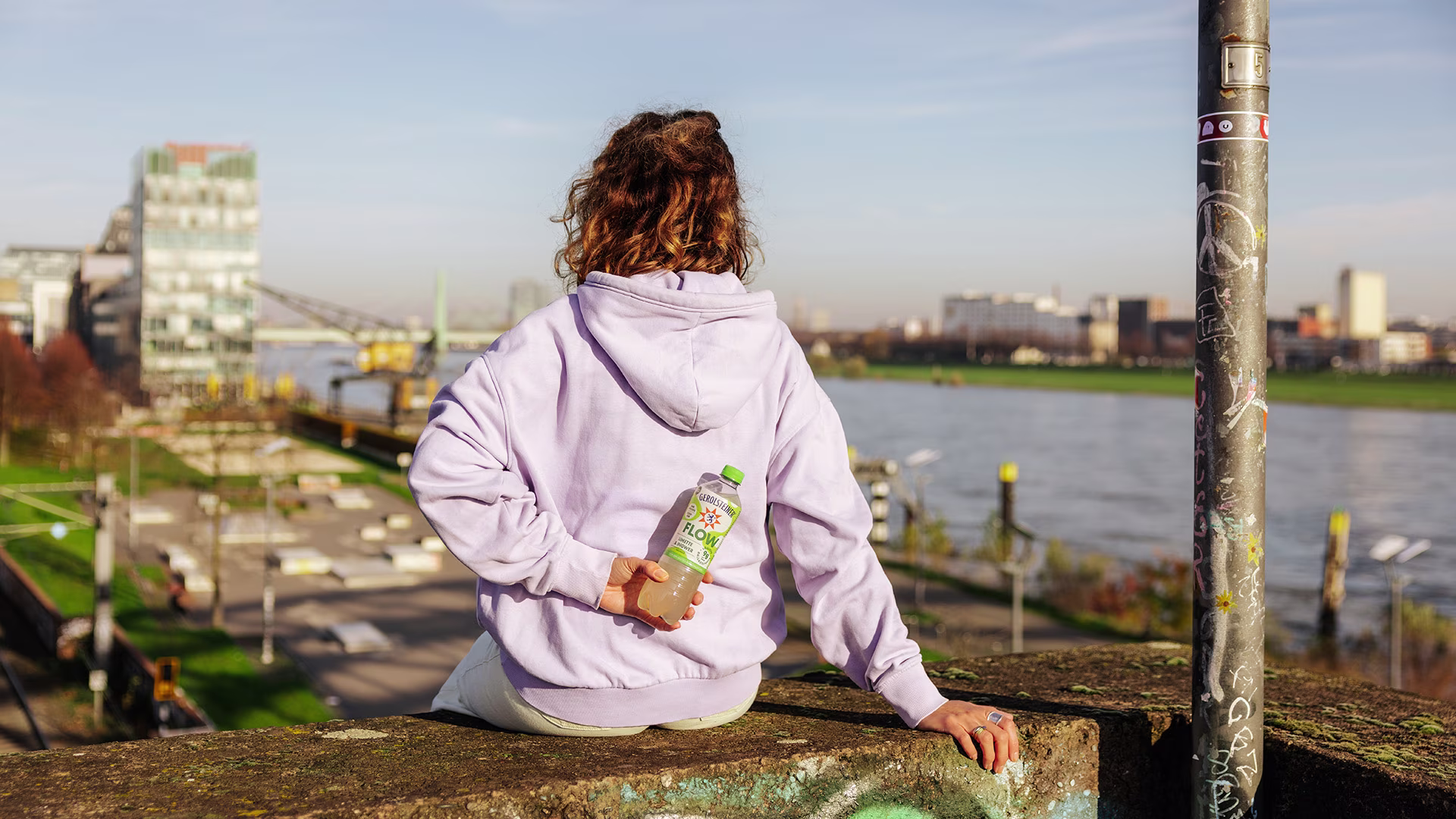 Eine Person sitzt mit dem Rücken zur Kamera auf einer Mauer mit Blick auf den Rhein und hält eine Flasche Gerolsteiner Flow Limette & Ingwer hinter dem Rücken. Die Szene wirkt entspannt und sonnig.