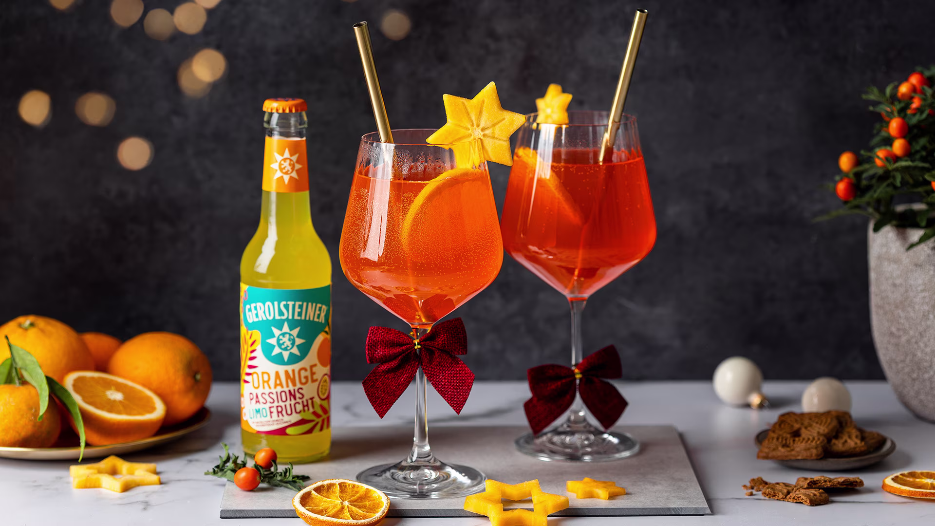 Zwei Gläser mit orangefarbenem Getränk, garniert mit Orangenscheiben und Sternfrucht, stehen neben einer Flasche Gerolsteiner Orange Passionsfrucht. Rote Schleifen, Orangen, Gebäck und Lichter sorgen für eine festlich-warme Atmosphäre.