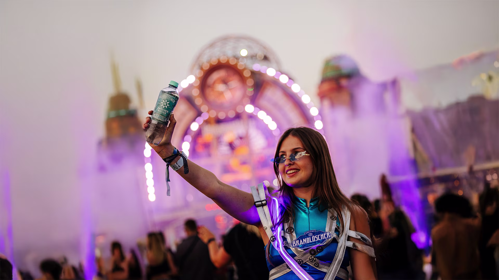 Junge Frau im futuristischen Outfit hält eine Flasche Gerolsteiner in die Höhe, vor einer Festivalbühne mit Lichteffekten und Menschenmenge bei Parookaville.
