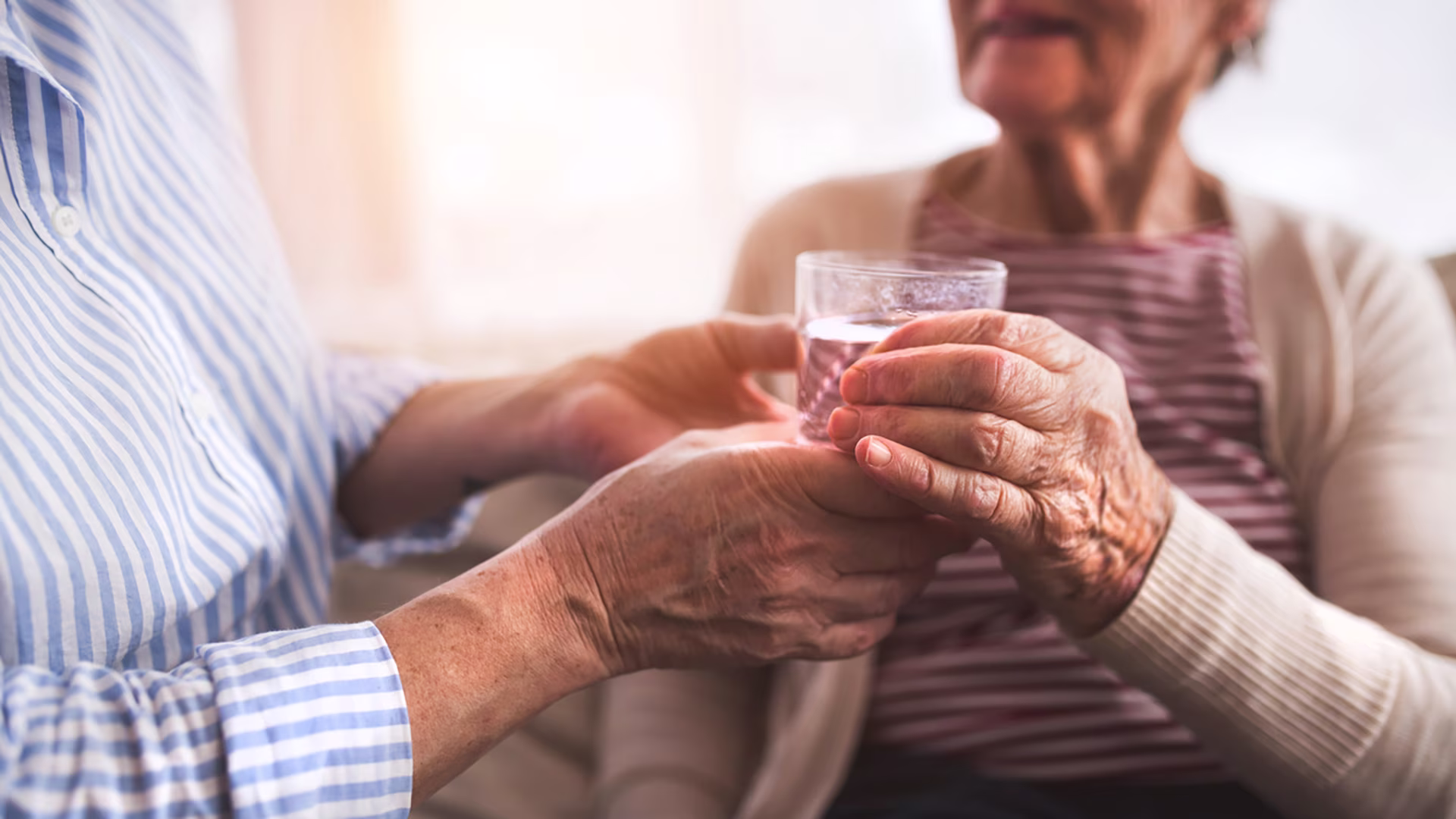 Ältere Frau hält ein Glas Wasser in den Händen, während eine andere Person sie dabei unterstützt.